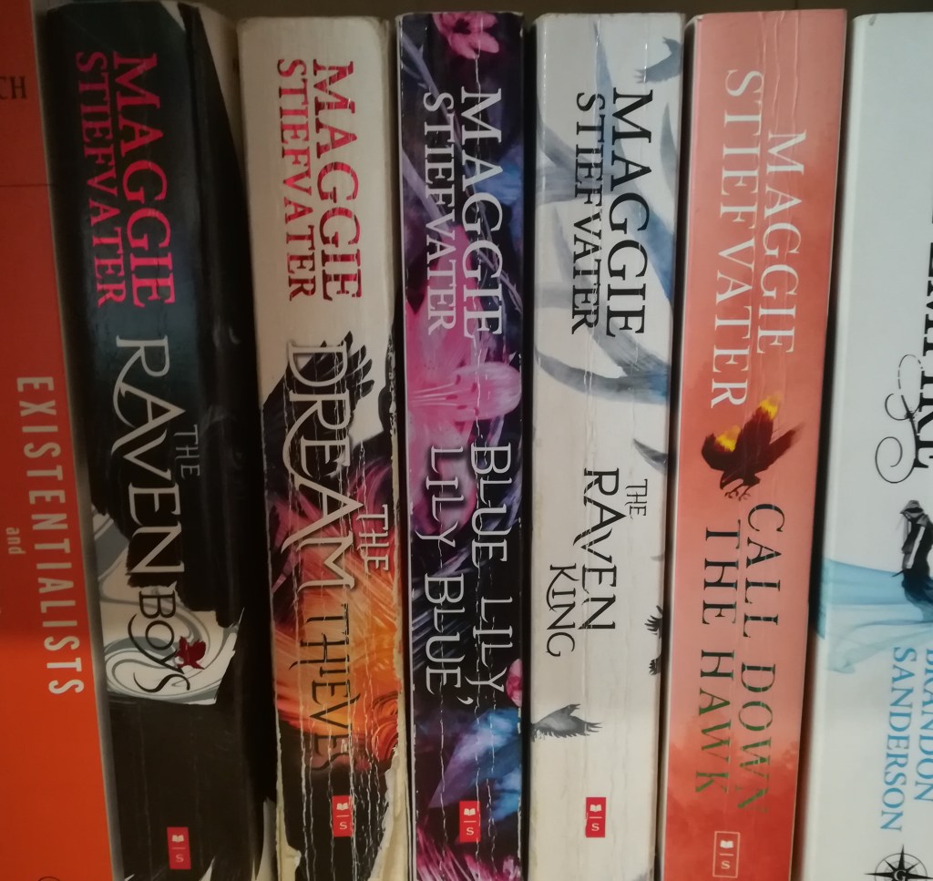 spine photograph of Maggie Stiefvater's UK editions of 'The Raven Cycle' series plus 'Call Down the Hawk. Partial showing of Iris Murdoch's 'Existentialists and Mystics' and Brandon Sanderson's 'The Final Empire', also in paperback.