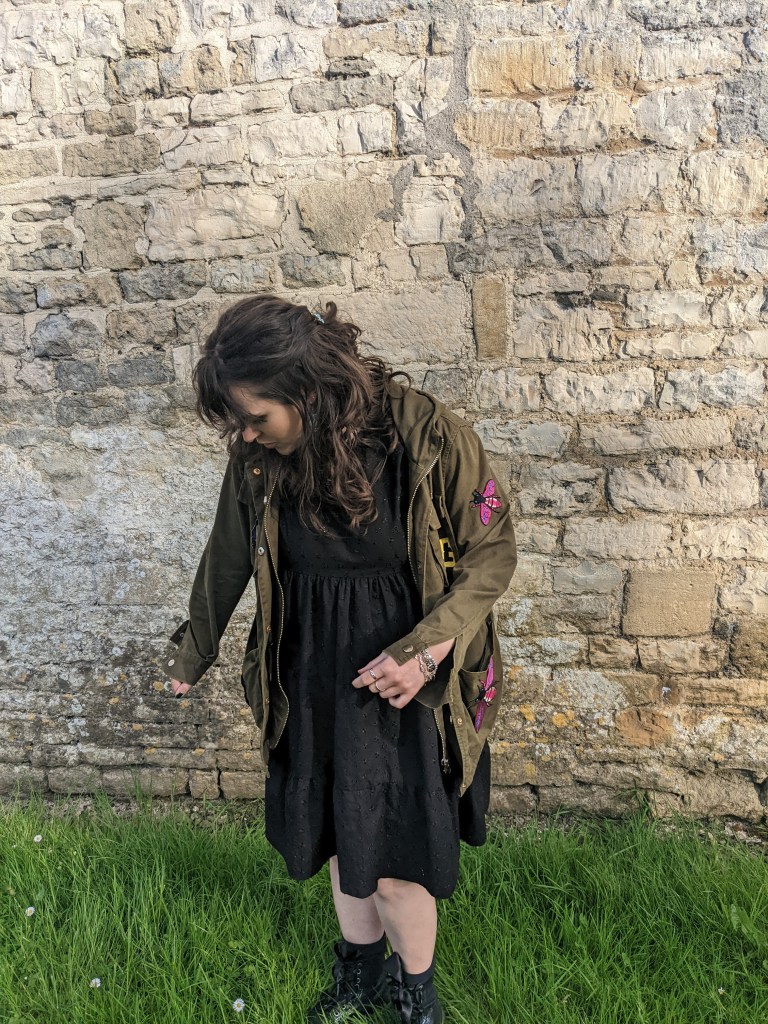short lady with dark hair, wearing a black dress and patch-covered khaki jacket
