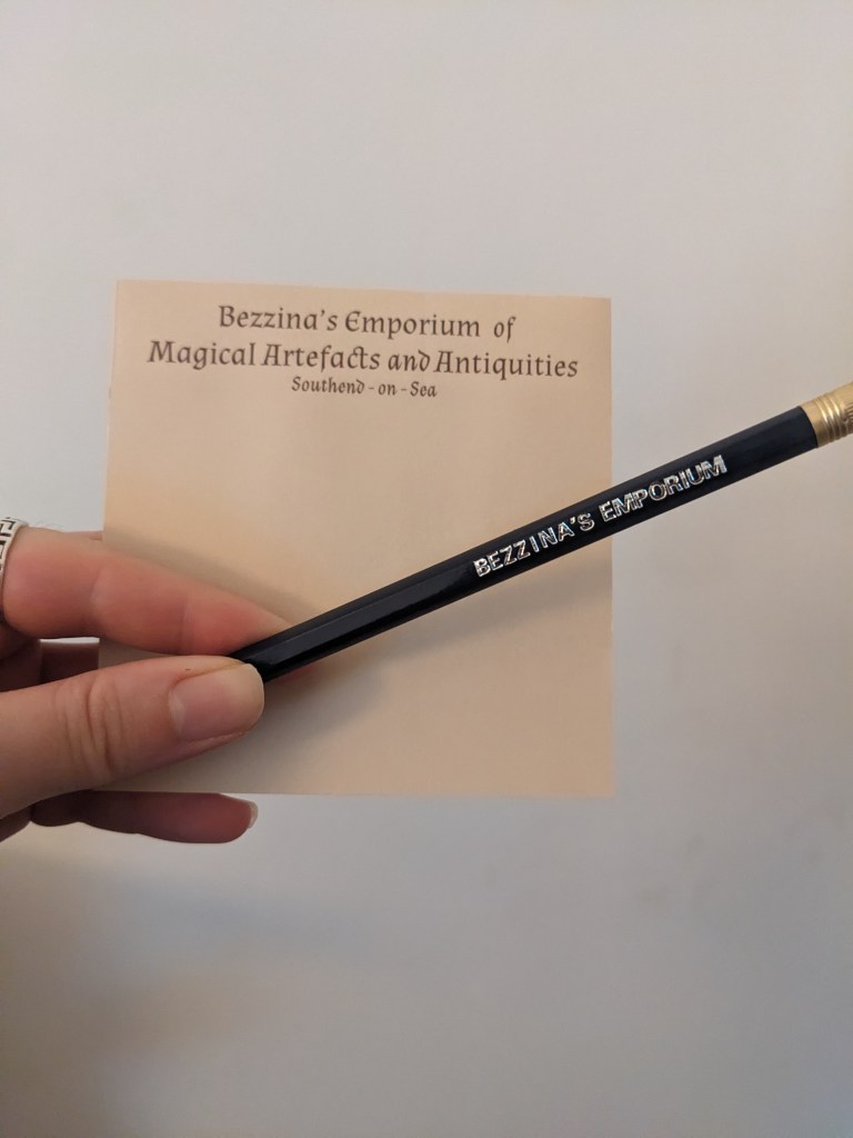 sticky note with brown writing reading 'Bezzina's Emporium of Magical Artefacts and Antiquities Southend-on-Sea' and blue and gold pencil with silver stamped 'Bezzina's Emporium' lettering