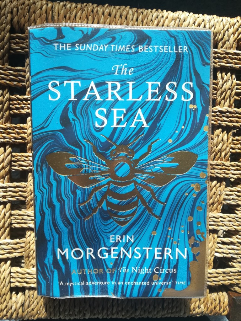 blue and gold paperback of The Starless Sea by Erin Morgenstern, in a plastic library cover, in a wicker stool in sunlight.
